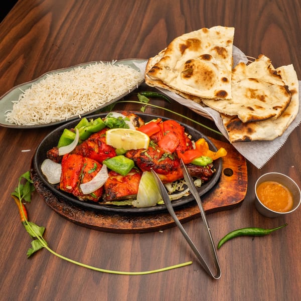 Chicken tikka sizzler with bell peppers, onions, lemon, rice, naan, and sauce on a wooden table.