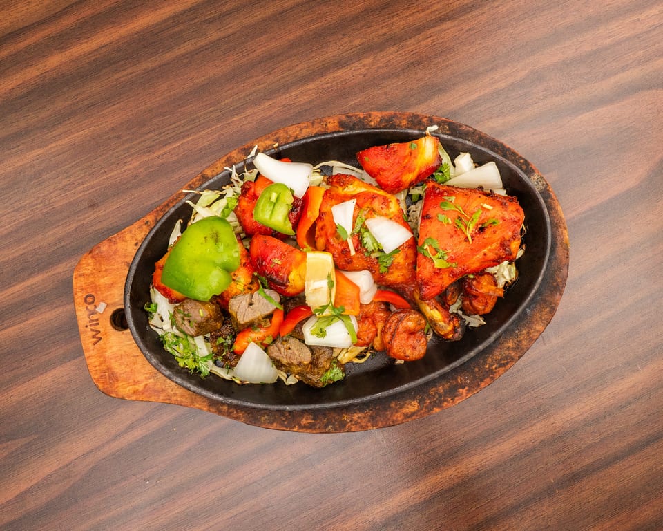 Sizzling platter of tandoori chicken, lamb, shrimp, and vegetables on a wooden table.