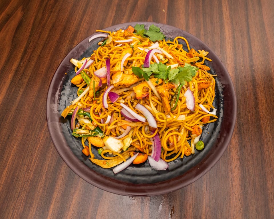 A black plate filled with chicken noodles, red onions, green chilies, and cilantro on a wooden table.