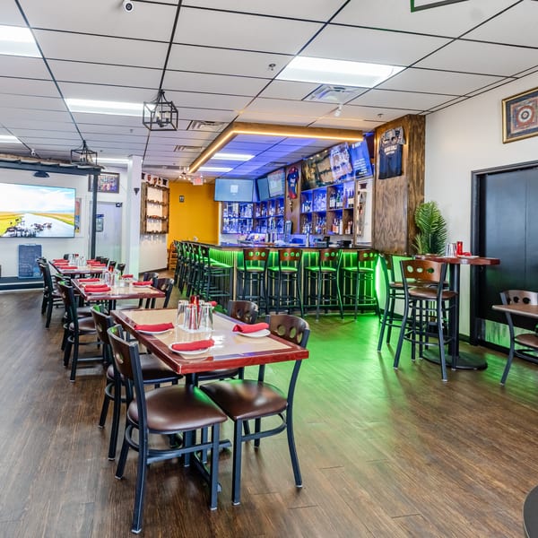 Restaurant interior with a long bar illuminated by green lights, tables with red napkins, and a large screen.
