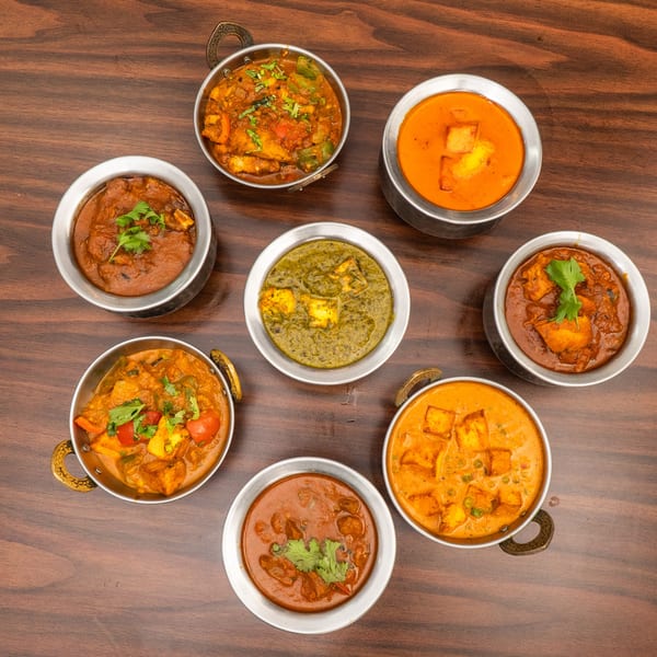 Seven bowls of various Indian curries, including paneer and vegetable dishes, arranged on a wooden table.