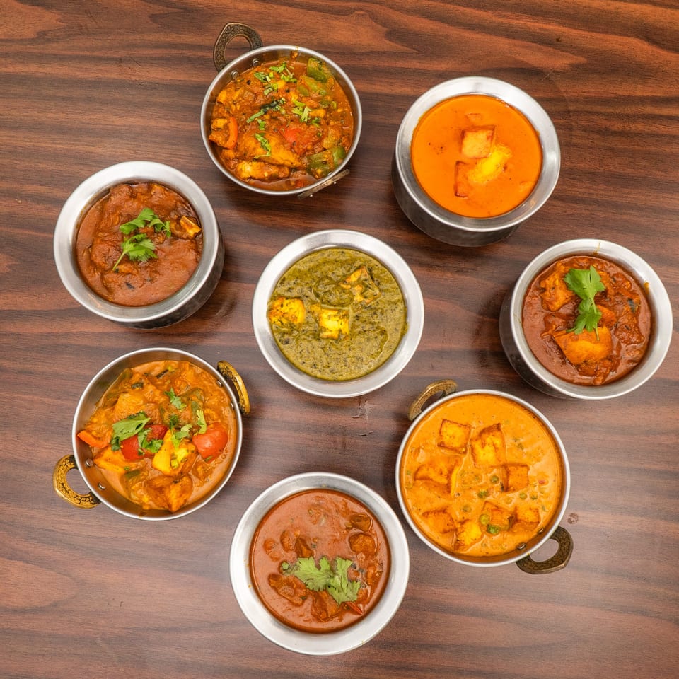Seven bowls of various Indian curries, including paneer and vegetable dishes, arranged on a wooden table.