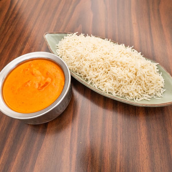 A metal bowl of orange curry and a green plate of white rice on a wooden table.