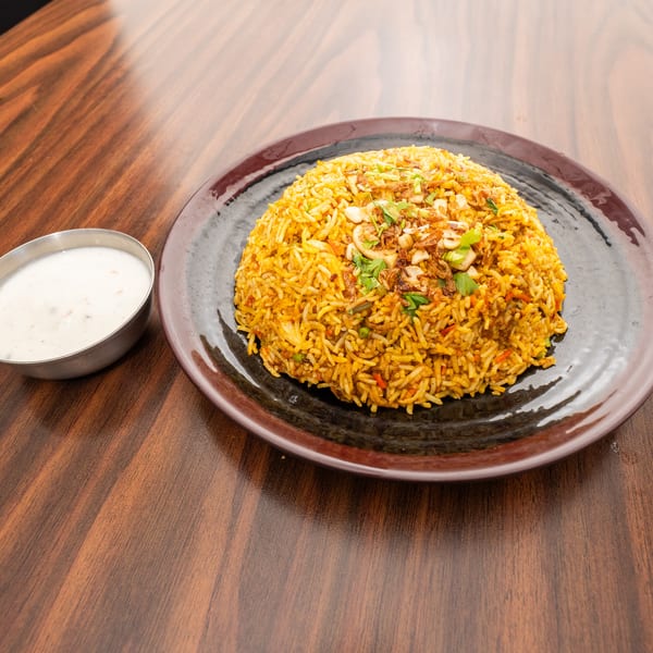 A mound of golden biryani rice with cashews and herbs, served with a side of white raita.