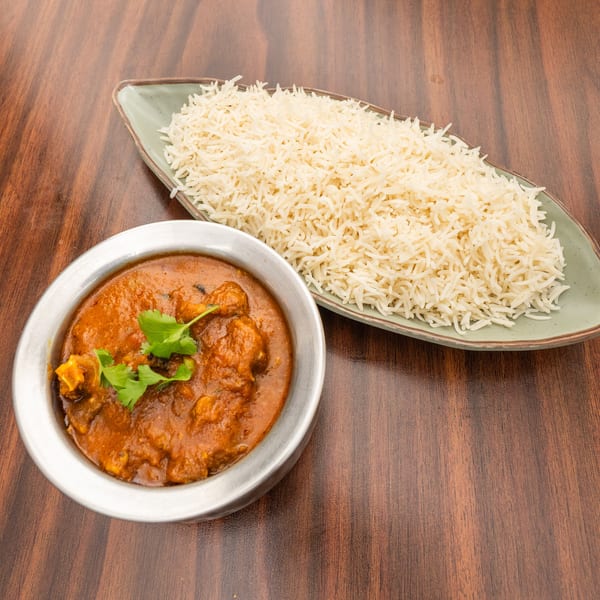 A bowl of chicken curry with cilantro garnish and a plate of white rice on a wooden table.