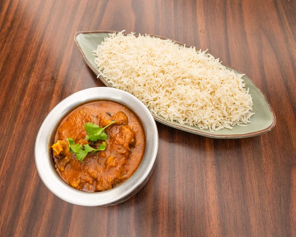 A bowl of chicken curry with cilantro garnish and a plate of white rice on a wooden table.