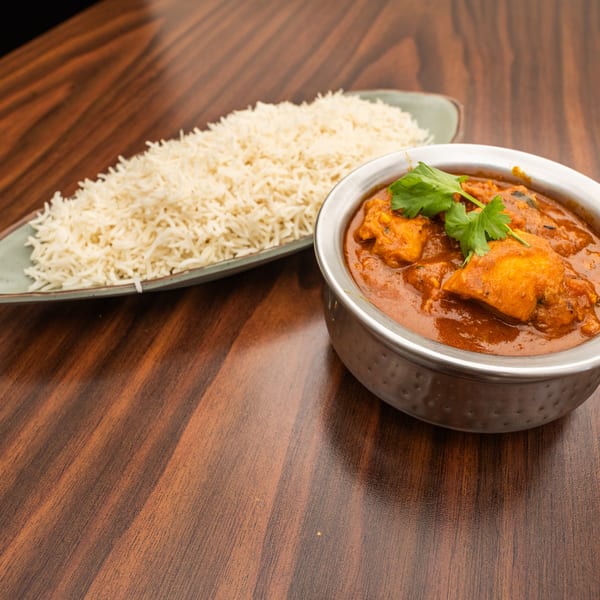 Chicken curry in a metal bowl with cilantro, served alongside white rice on a wooden table.