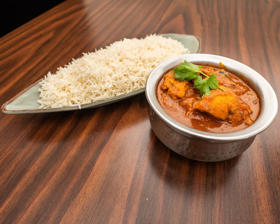 Chicken curry in a metal bowl with cilantro, served alongside white rice on a wooden table.