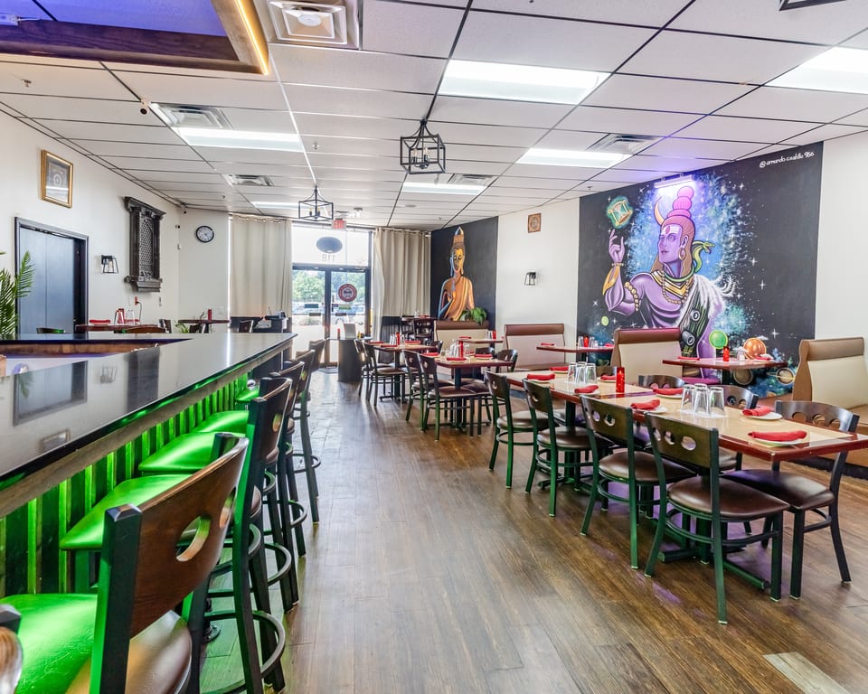 Restaurant interior with a long bar and green stools, dining tables, and colorful murals on the walls.