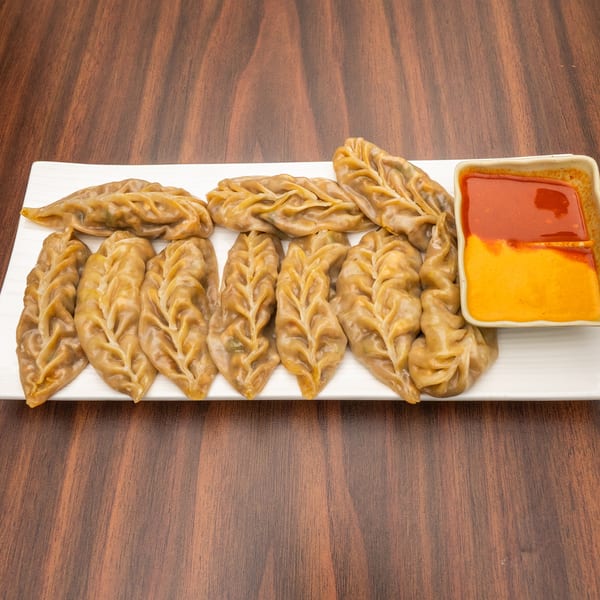 A white rectangular plate holds a row of steamed momos with a side of red and orange dipping sauce.