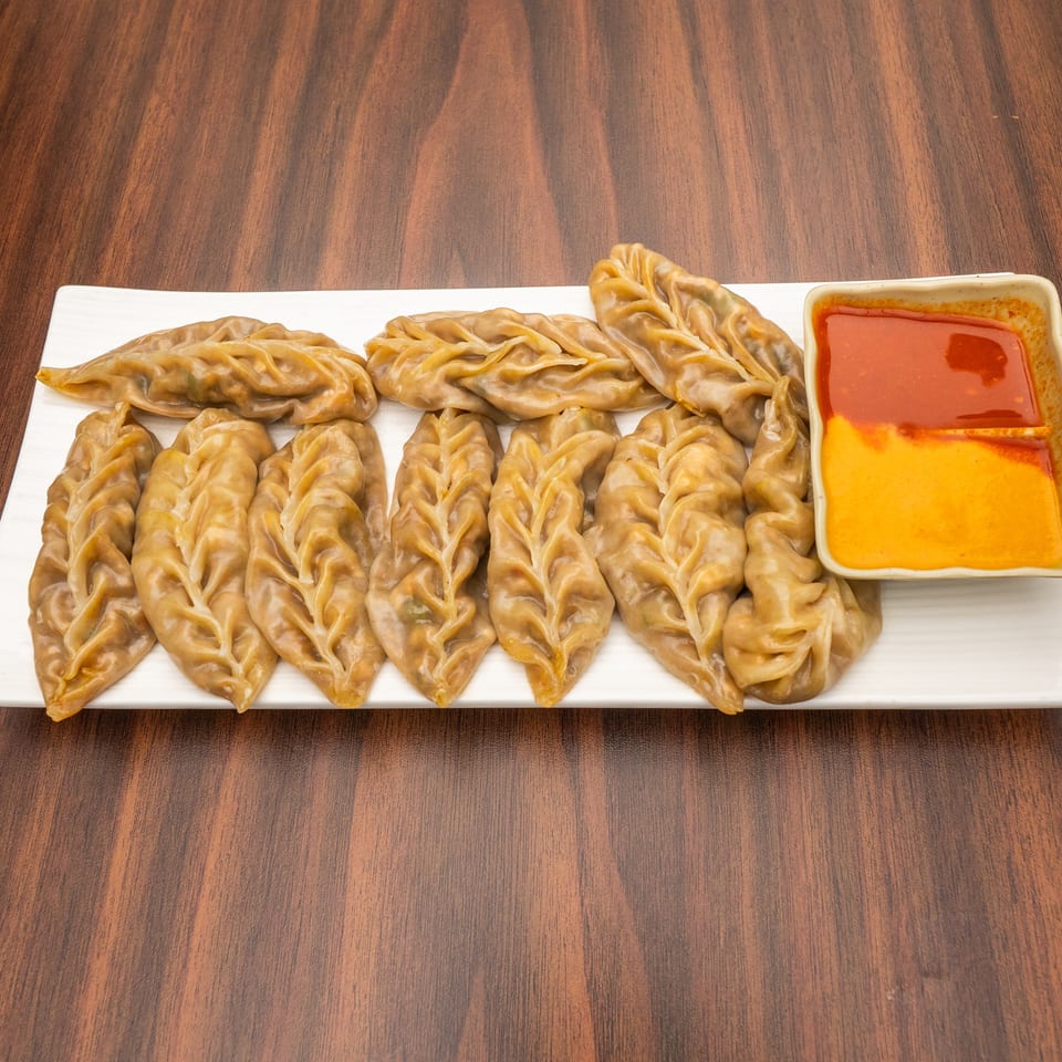 A white rectangular plate holds a row of steamed momos with a side of red and orange dipping sauce.