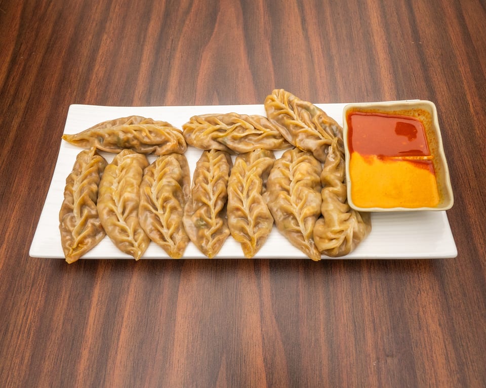 A white rectangular plate holds a row of steamed momos with a side of red and orange dipping sauce.
