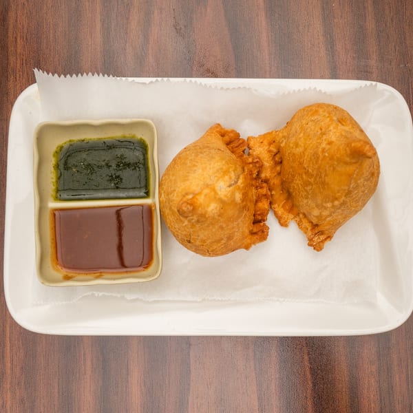 Two golden-brown samosas and a divided dish of green and brown chutneys on a white plate.