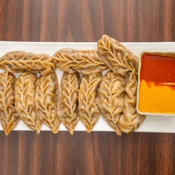 A white rectangular plate of steamed momos with intricate leaf-like pleats, served with two dipping sauces.