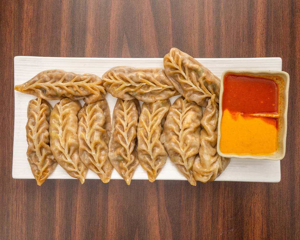 A white rectangular plate of steamed momos with intricate leaf-like pleats, served with two dipping sauces.