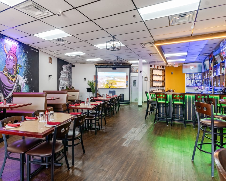 A restaurant interior with dining tables, booths, a bar with green lighting, and wall murals.