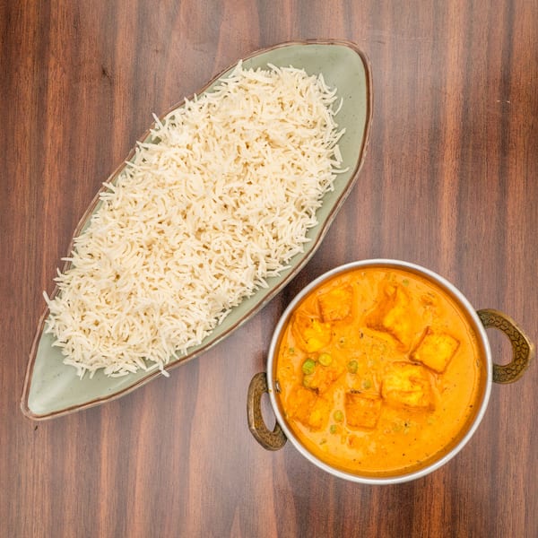 Overhead view of paneer butter masala with peas in a metal bowl and basmati rice on a green plate.